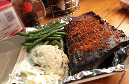 A plate featuring a seasoned slab of meat, green beans, and creamy mashed potatoes, served in a takeout container.