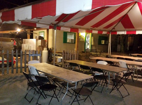 A tent with red and white stripes covers several empty tables and chairs outside a building at night.