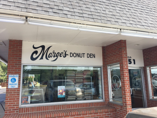 Sign for "Marge's Donut Den" on a brick building, with a window displaying colorful posters and a parking sign.