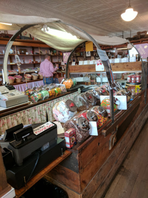 A rustic candy shop display with jars of colorful sweets and a wooden counter, featuring a cash register in the foreground.