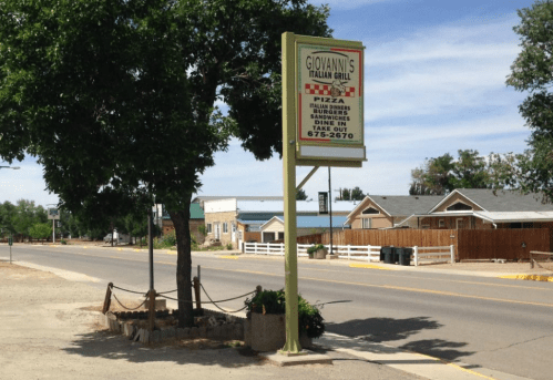 Sign for Giovanni's Italian Grill, featuring pizza and sandwiches, located on a quiet street with trees and buildings nearby.