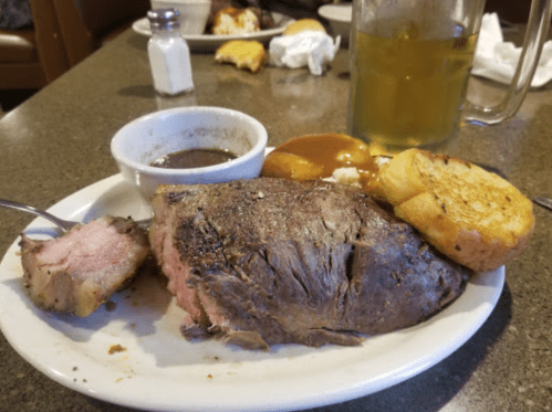 A plate with a slice of roast beef, gravy, a side of sauce, garlic bread, and a drink in the background.