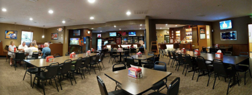 A spacious restaurant interior with tables, chairs, and a bar area, featuring patrons and multiple TVs on the walls.
