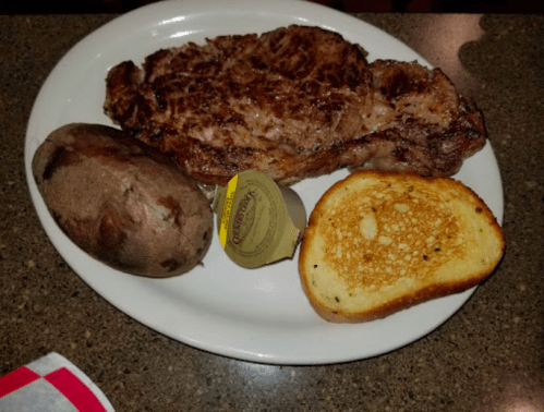 A plate with a grilled steak, baked potato, garlic butter, and toasted bread.