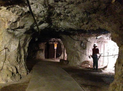 A dimly lit underground mine tunnel with rocky walls and a person standing near a cable.