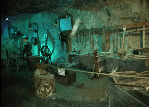 A dimly lit underground workshop with tools, machinery, and a figure working in a cave-like setting.