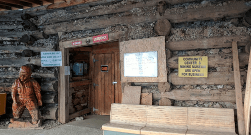 Entrance to a rustic building with a wooden exterior, featuring a sign and a carved figure. Community center open for business.