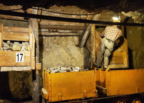 A miner loads rocks into a cart inside a dimly lit underground mine. Wooden structures and tools are visible.