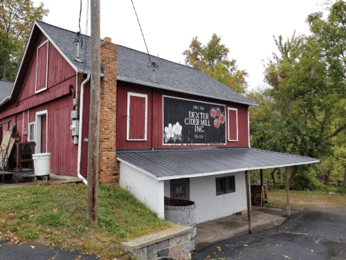 Red wooden building with a sign for Dexter Cider Mill, surrounded by trees and a cloudy sky.