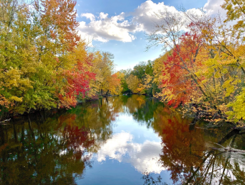 A serene river reflects vibrant autumn foliage in shades of red, orange, and yellow under a partly cloudy sky.