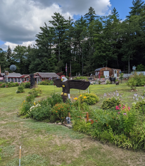 A garden with colorful flowers and a wooden eagle sculpture, surrounded by trees and rustic buildings in the background.