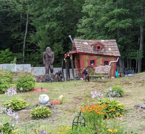 A whimsical garden scene featuring a small red house, a Bigfoot statue, and various colorful flowers.