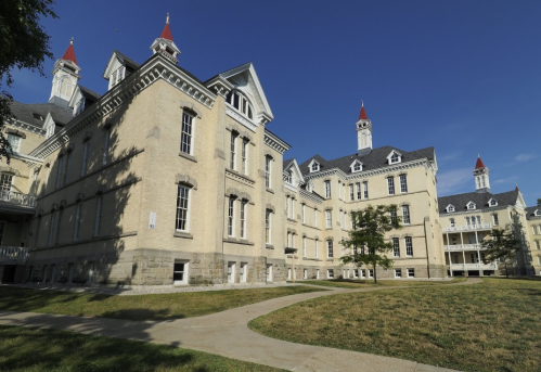 Historic building with a light-colored facade, red-tipped spires, and a grassy area under a clear blue sky.