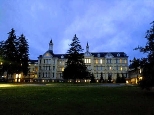 Historic building at dusk, illuminated windows, surrounded by trees and a grassy area.