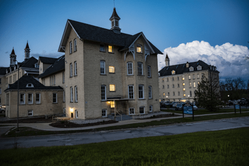 Historic buildings at dusk, featuring illuminated windows and a cloudy sky in the background.