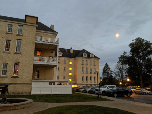 Historic brick buildings at dusk, with a full moon and parked cars in the foreground. Soft lighting from windows.