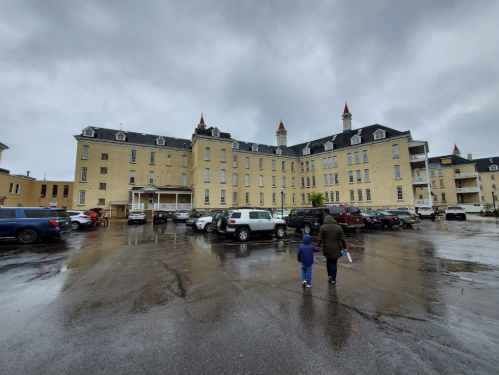 A child and adult walk towards a large, historic building on a rainy day, with parked cars in the foreground.