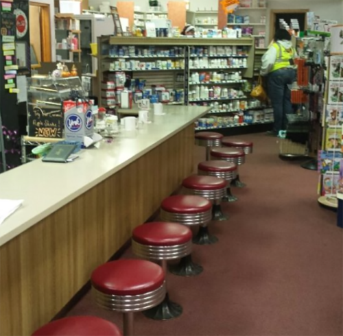 A retro diner interior with a long counter and red stools, shelves filled with products, and a person in a reflective vest.