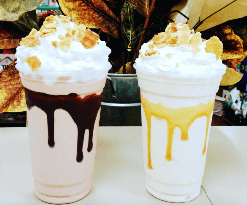 Two milkshake cups topped with whipped cream and drizzled with chocolate and caramel, garnished with crushed cereal.