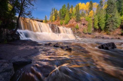 A scenic waterfall surrounded by autumn foliage and lush green trees, with flowing water and a clear blue sky.