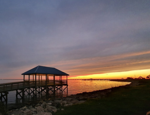 A wooden pier extends over calm waters at sunset, with vibrant orange and purple hues in the sky.