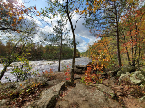 A scenic view of a river surrounded by colorful autumn trees and rocky terrain under a partly cloudy sky.