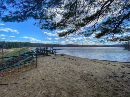 A sandy riverbank with a wooden platform, surrounded by trees and a clear blue sky with scattered clouds.