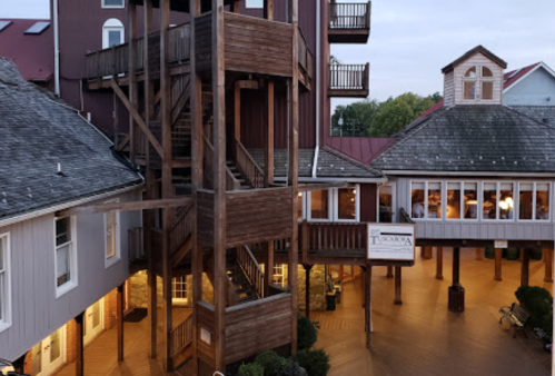 A wooden structure with stairs stands in a courtyard surrounded by buildings, illuminated in the evening light.