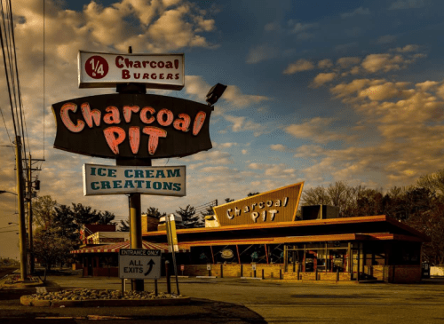Sign for Charcoal Pit featuring "Charcoal Burgers" and "Ice Cream Creations" with a retro-style building in the background.