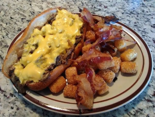 A plate featuring a cheesy steak sandwich with bacon and a side of crispy tater tots.