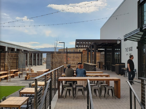 Outdoor dining area with wooden tables, string lights, and a modern building in the background under a blue sky.