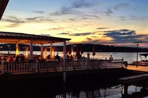 A waterfront restaurant at sunset, with diners enjoying the view and colorful clouds reflecting on the water.