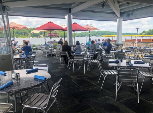Outdoor dining area with metal tables and chairs, umbrellas, and a view of a lake, filled with patrons enjoying their meals.