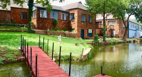 A rustic stone house beside a calm pond, with a wooden dock and lush greenery surrounding the area.