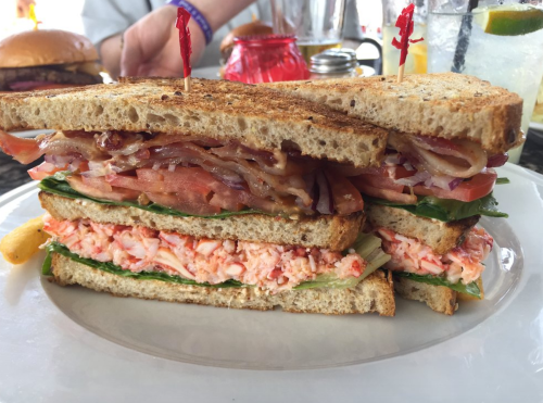 A layered sandwich with lobster, lettuce, tomatoes, and bacon on toasted bread, garnished with toothpicks.
