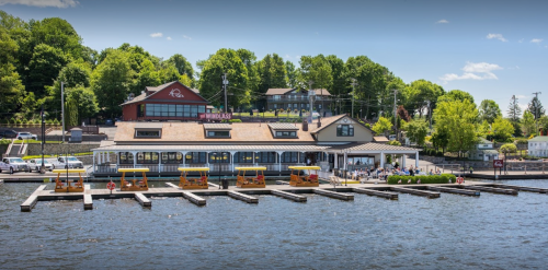 A waterfront building with a restaurant and docked boats, surrounded by green trees and a clear blue sky.