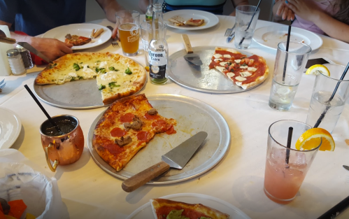 A table with various pizzas, drinks, and plates, featuring slices of pepperoni, white pizza, and a cocktail.