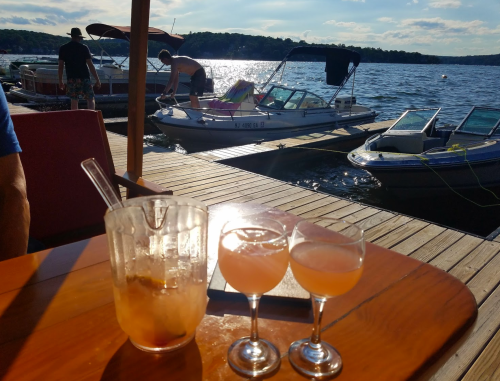 Two cocktails on a table with boats docked on a sunny lake in the background.