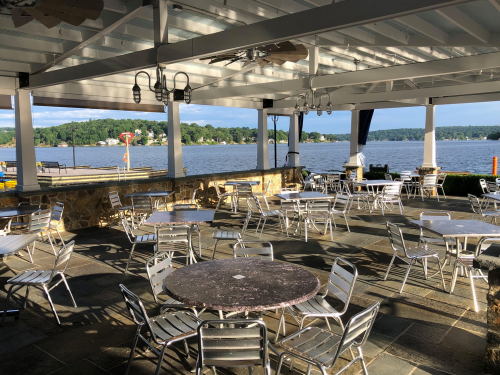 Outdoor dining area with metal tables and chairs overlooking a lake, under a covered patio with a scenic view.