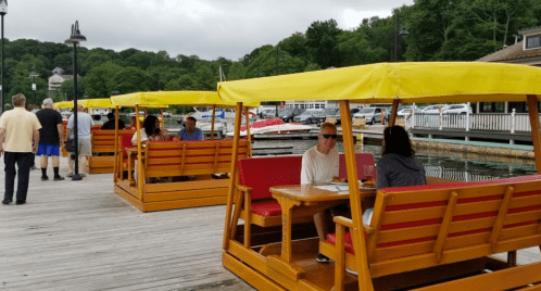 People dining at outdoor tables with yellow canopies by a marina, surrounded by boats and greenery.