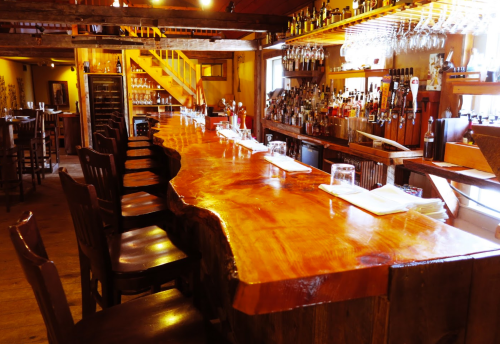 A rustic bar with a polished wooden counter, high stools, and shelves stocked with various bottles in a warm, inviting setting.