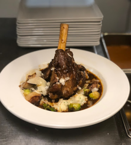 A plated dish featuring a large lamb shank on creamy mashed potatoes, garnished with Brussels sprouts and rich sauce.