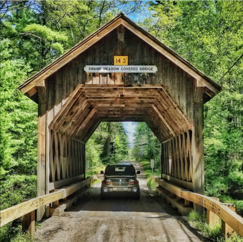 A car drives through a wooden covered bridge surrounded by lush green trees. A sign indicates the bridge height.