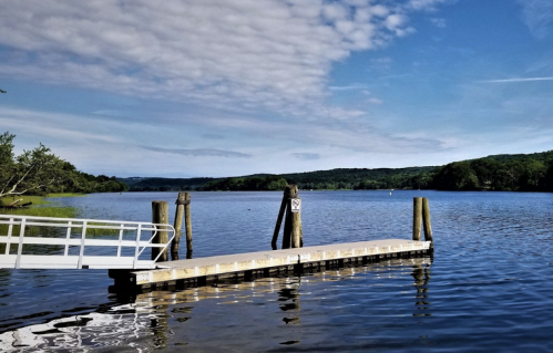 A serene lake view with a wooden dock extending into calm waters, surrounded by green hills and a blue sky.