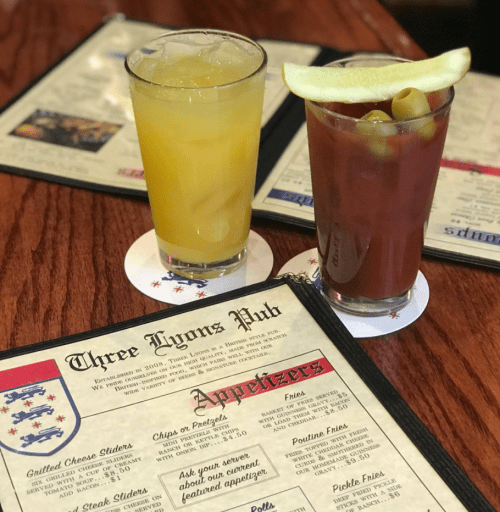 Two drinks on a table: a yellow cocktail and a Bloody Mary, with a menu from Three Lyons Pub in the background.