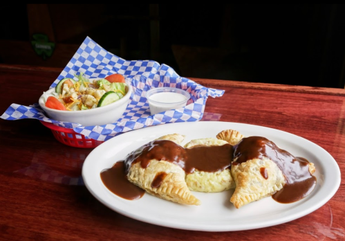 A plate of two stuffed pastries with brown sauce, alongside a salad in a basket with tomatoes and cucumbers.