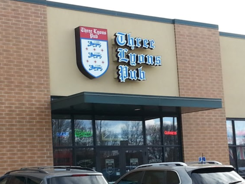 Exterior of Three Lyons Pub, featuring a sign with the pub's name and a brick facade. Cars parked in front.