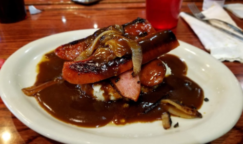 A plate of sliced sausage topped with grilled onions, served over mashed potatoes and covered in brown gravy.