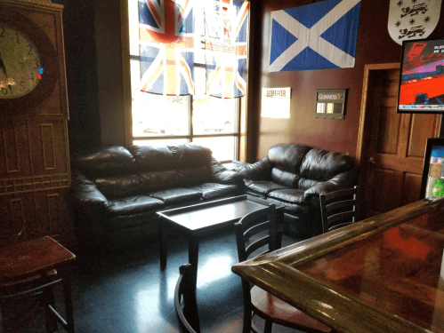 A cozy pub interior featuring leather sofas, a coffee table, and flags hanging on the walls.