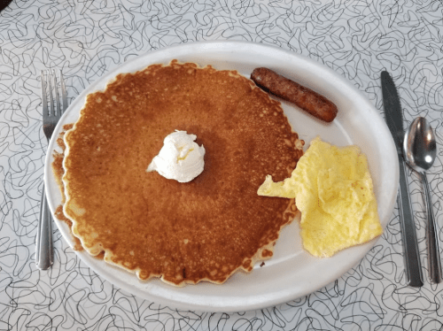 A plate with a large pancake topped with whipped cream, a sausage link, and scrambled eggs on a patterned table.